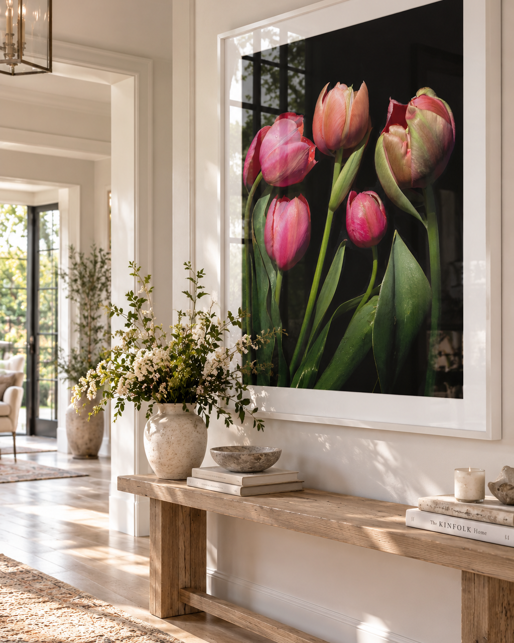 A large framed wall art print of Fine Romance, featuring pink tulips on a dark background on a white frame, displayed in a bright, modern entryway above a wooden bench with decorative items.