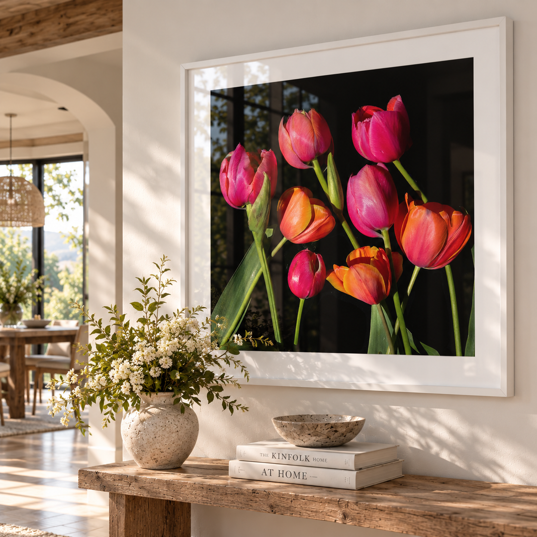 A framed wall art print featuring a cluster of pink and orange tulips on a dark background, displayed above a rustic wooden console table with a vase of white flowers and stacked books in a bright, modern interior.