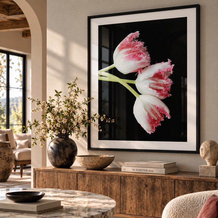 A framed floral wall art print hangs above a wooden console in a styled living room. The artwork shows three tulips with white petals and pink edges against a black background, enclosed in a black frame with a white mat.