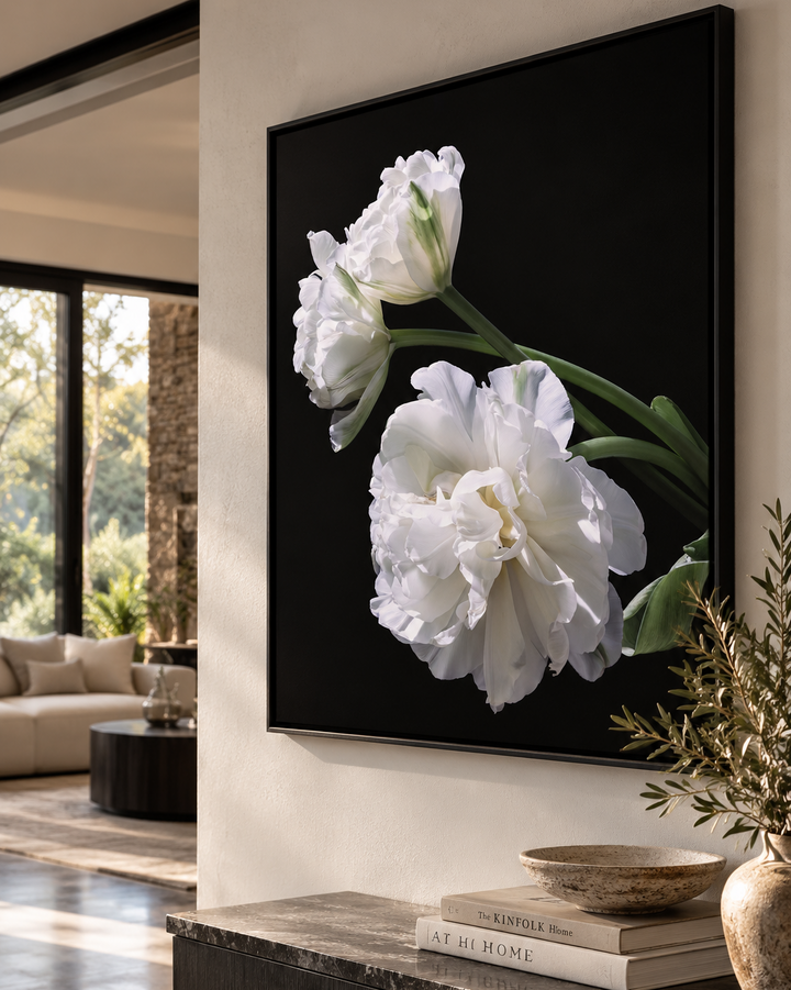 A framed black canvas wall art print featuring large white Tulip flowers with green stems, displayed above a console table in a modern living room setting.