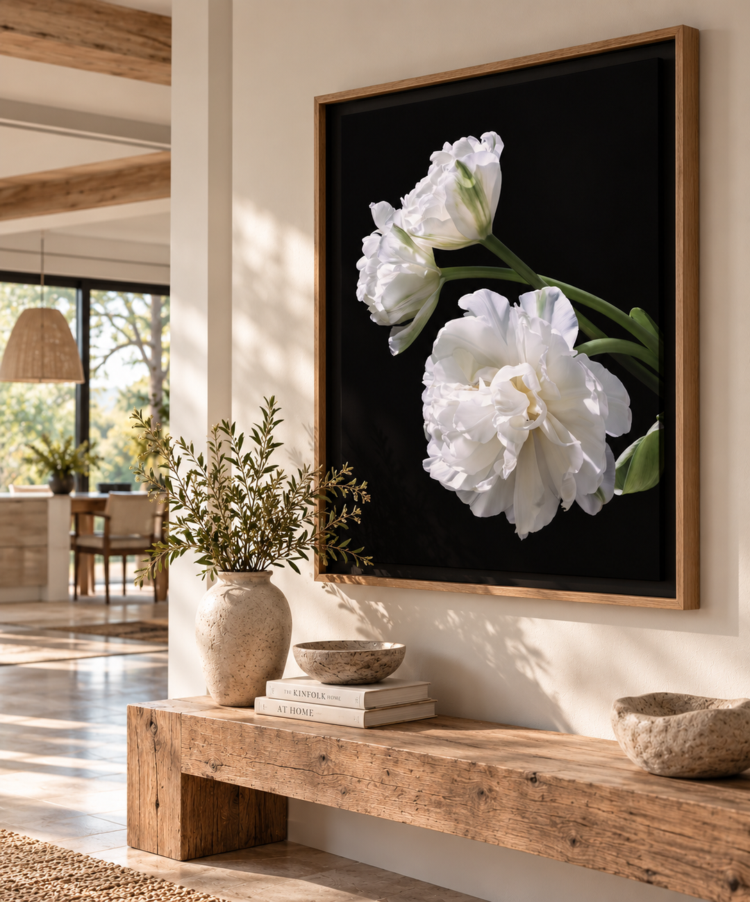A large framed wall art print featuring white tulips on a black background, hung above a rustic wooden bench in a modern, sunlit interior with beige walls and decorative vases.